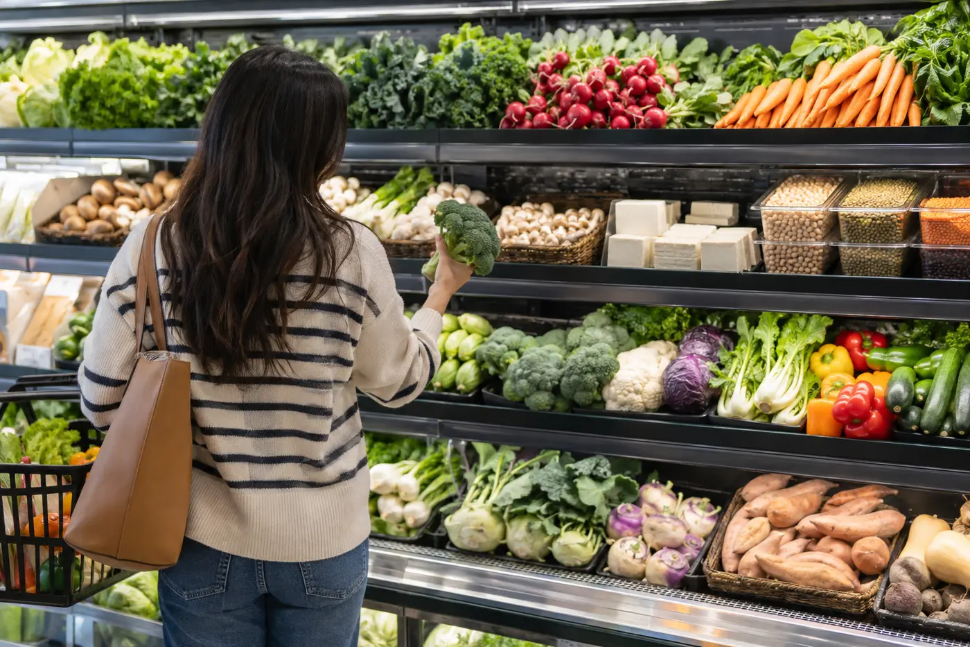 A shopper choosing broccoli beside vegetables, legumes and tofu in a grocery aisle.